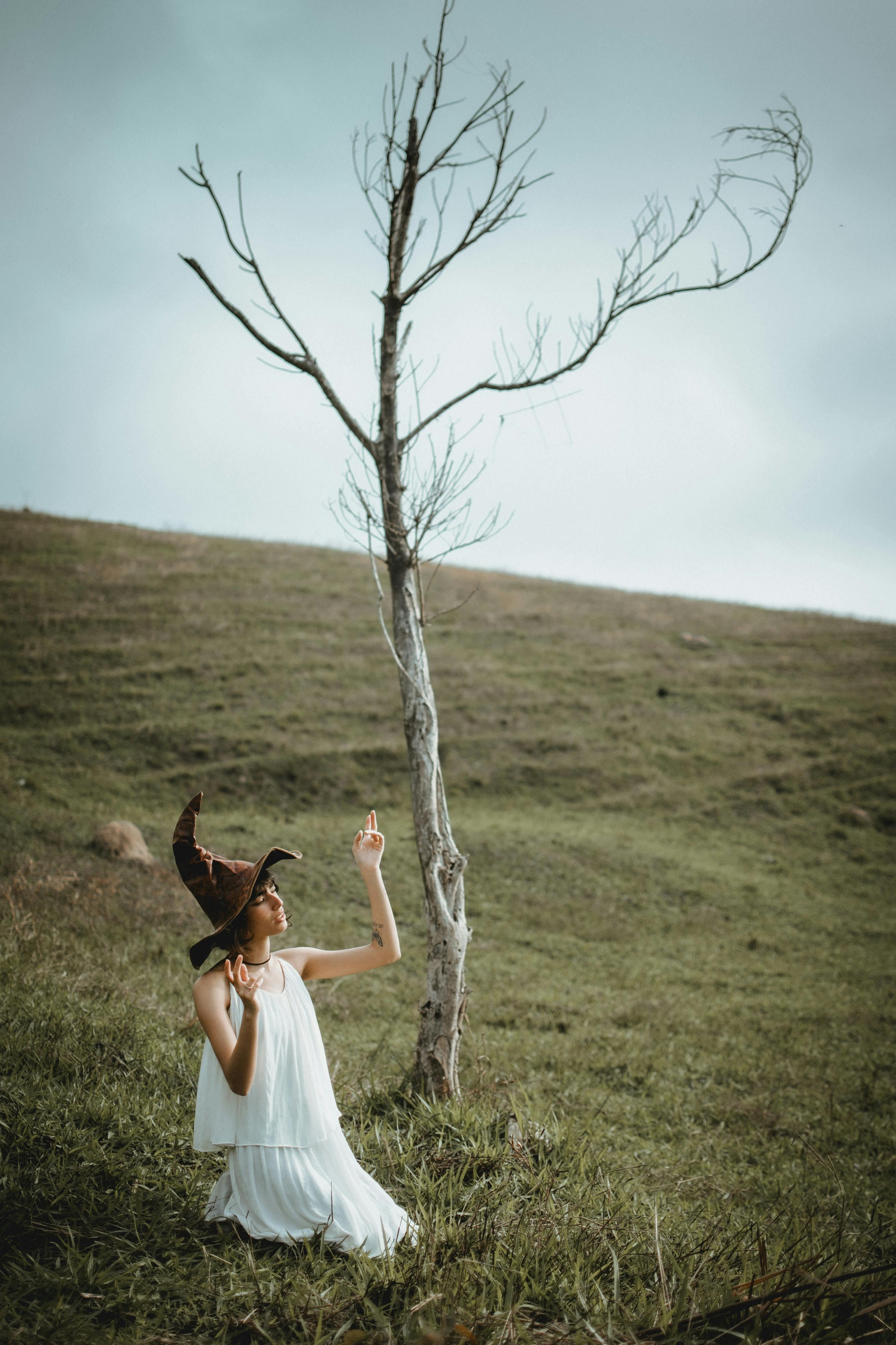 Woman in white dress kneeling beside leafless tree, wearing witch hat. Mystical ambiance on grassy hill.