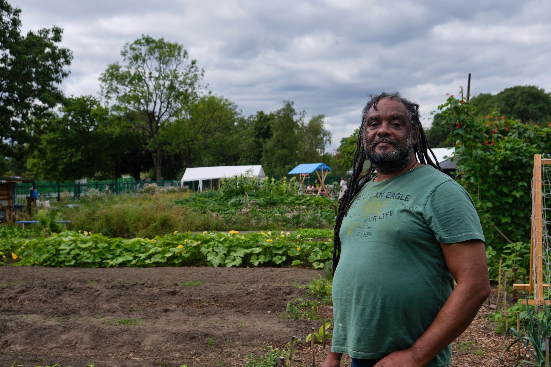 Positive unshaven African American male farmer with dreadlocks on head standing in garden of rural village