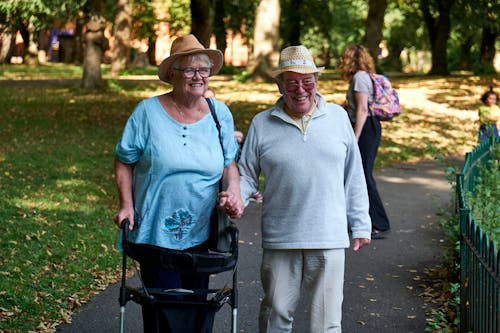 Cheerful elderly couple in casual clothes and straw hats walking on lawn of park in sunny day