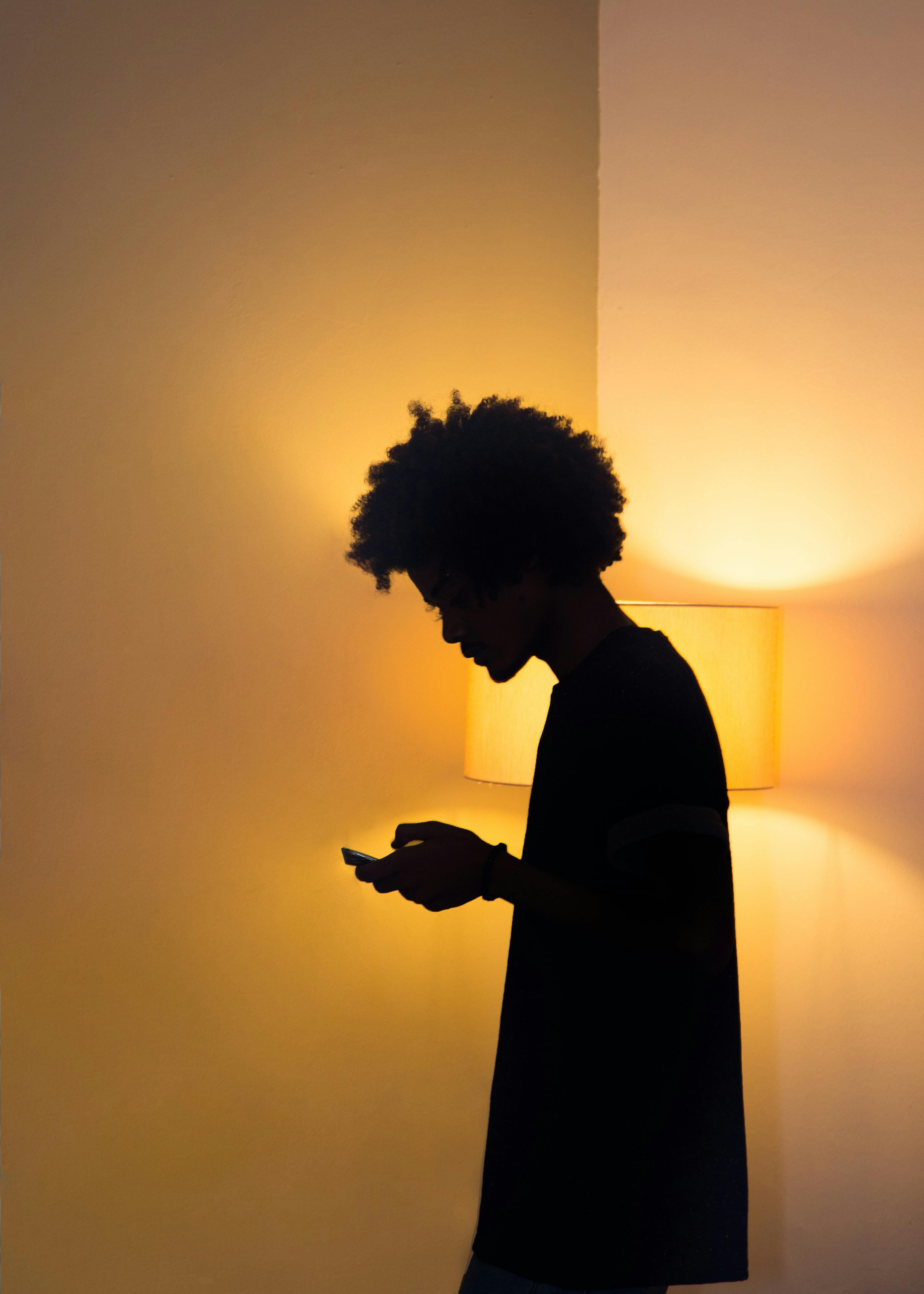 Silhouette of a person with afro hair using a smartphone in dim lighting, side view.