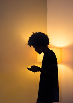 Silhouette of a person with afro hair using a smartphone in dim lighting, side view.