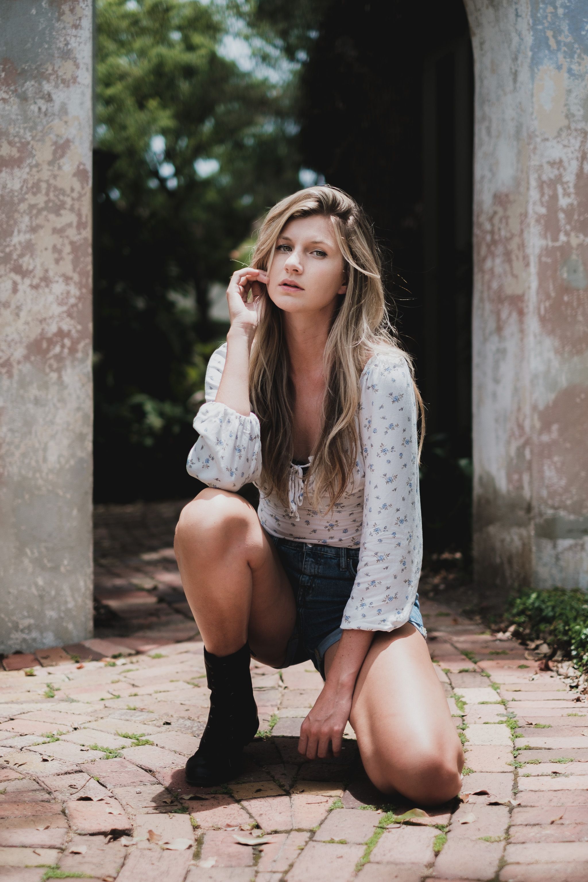Portrait of a woman in denim shorts and white blouse, kneeling on a brick path.