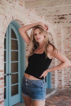 Young woman in casual attire poses by a rustic brick wall with blue doors.