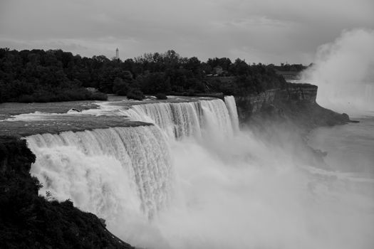 Black and white scenic view of Niagara Falls with mist and trees in the background.