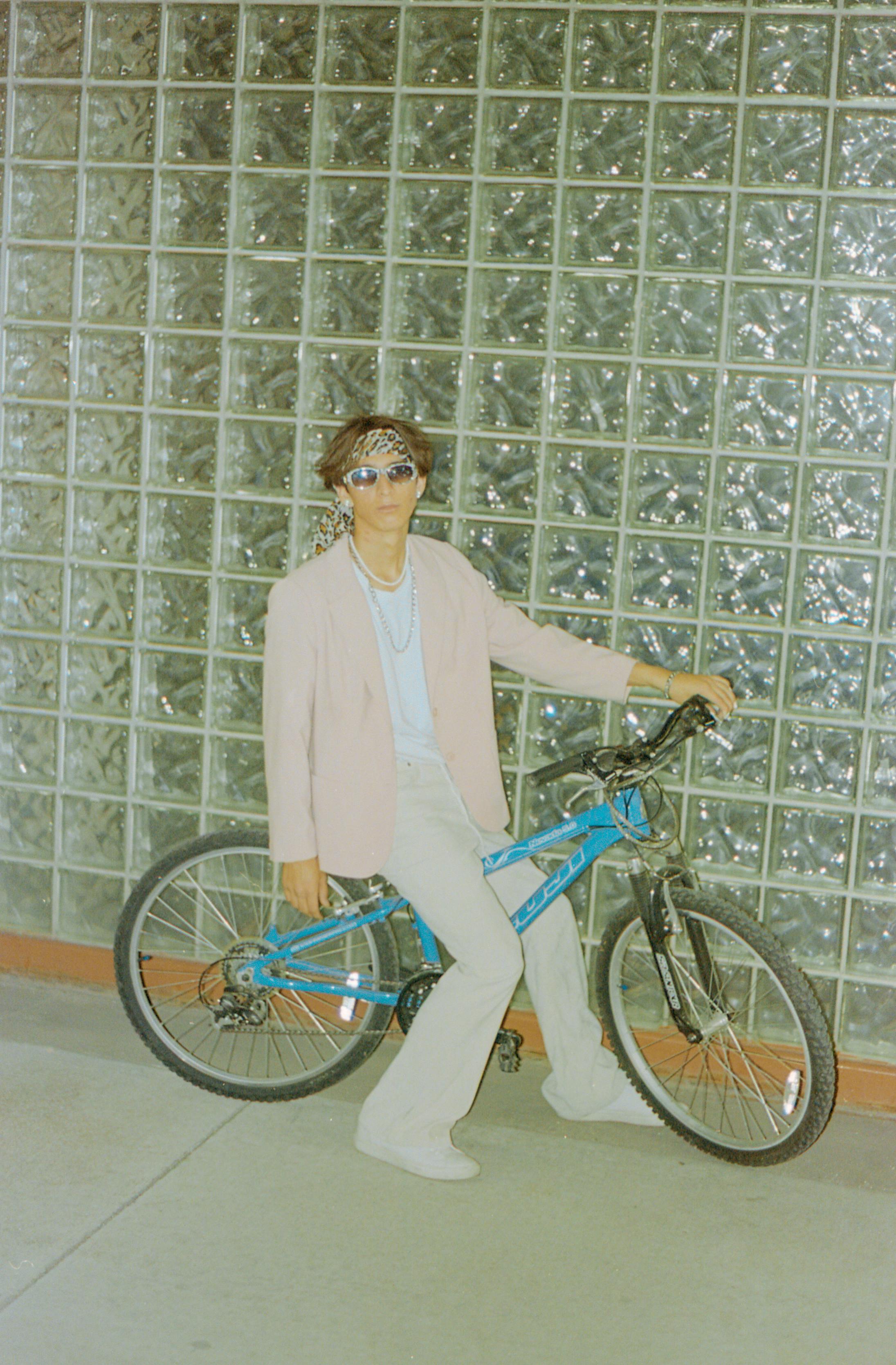 Stylish young adult seated on a bicycle against a glass wall, wearing sunglasses.