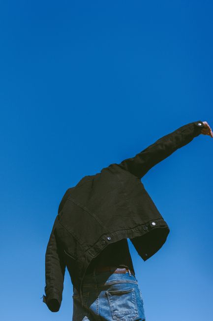 A person wearing denim and a black jacket against a bright blue sky, arms raised in motion.