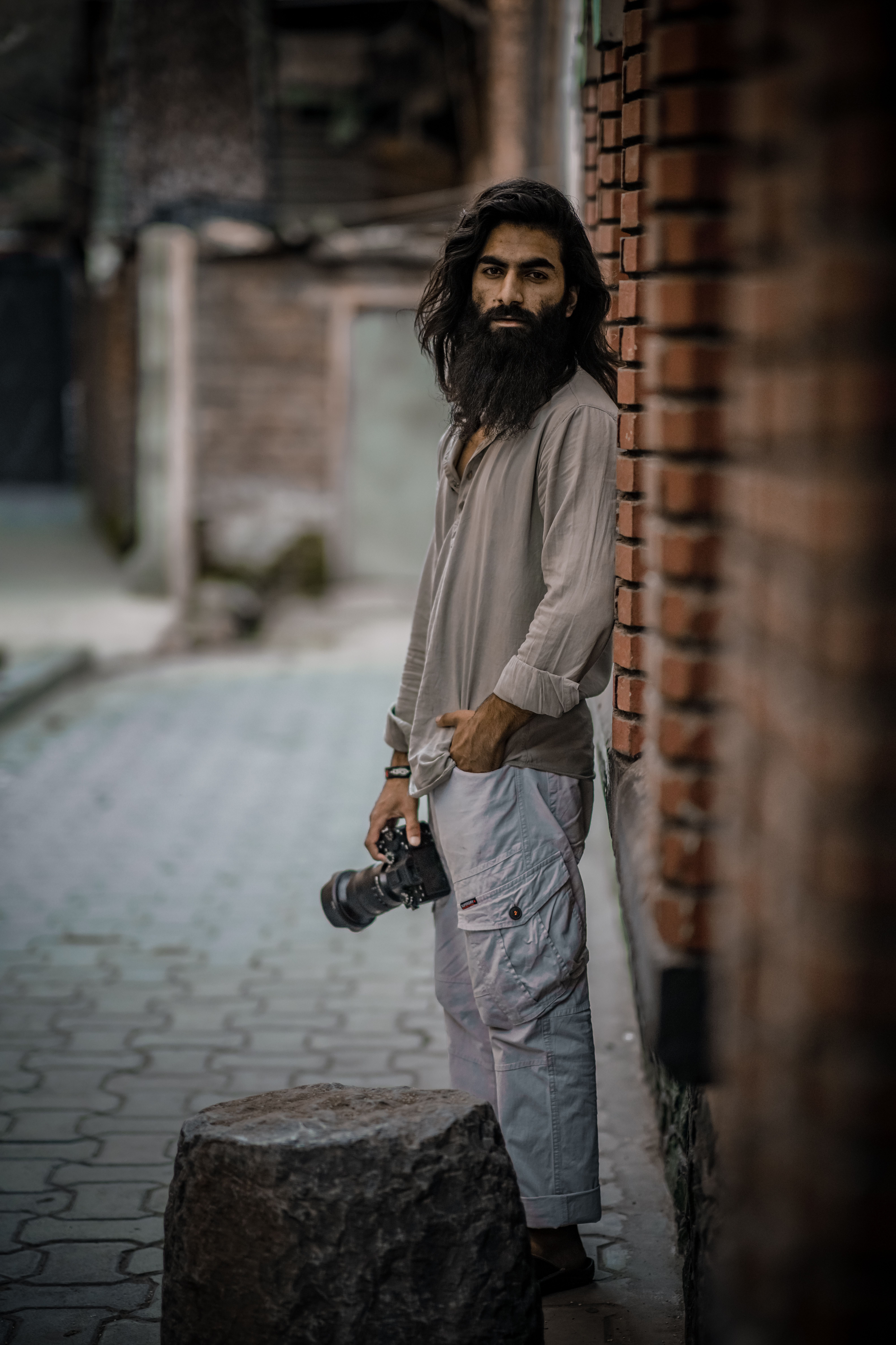 A bearded photographer poses outdoors with a camera in hand against a brick wall.