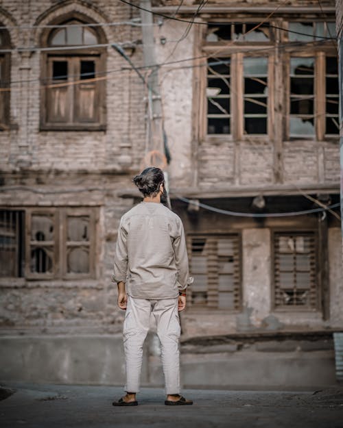 Man in Gray Long Sleeve Shirt and White Pants Standing Near Brick Building