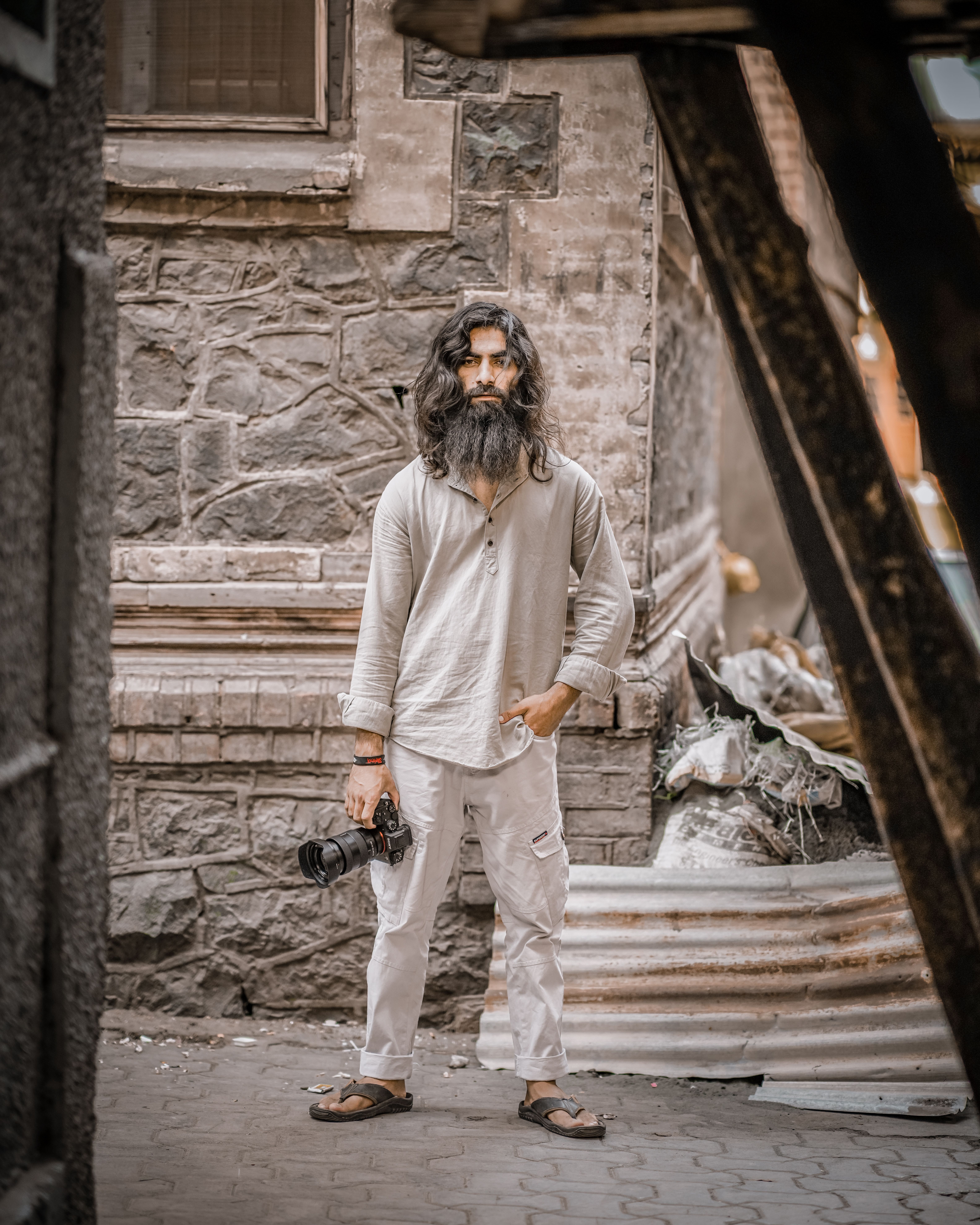 Bearded man with a DSLR camera posing against a rustic stone wall.