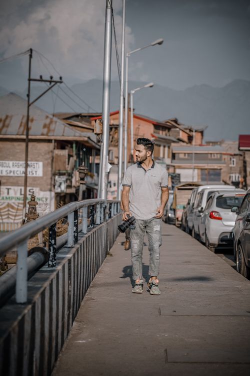Free Young man walking on a mountain town sidewalk holding a camera amidst a row of parked cars. Stock Photo