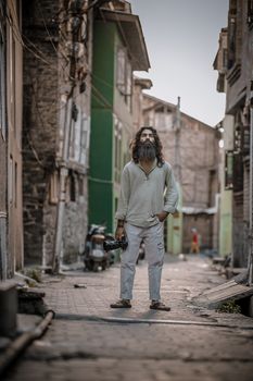 Portrait of a bearded man holding a camera in a rustic alley in JK, surrounded by vintage buildings.