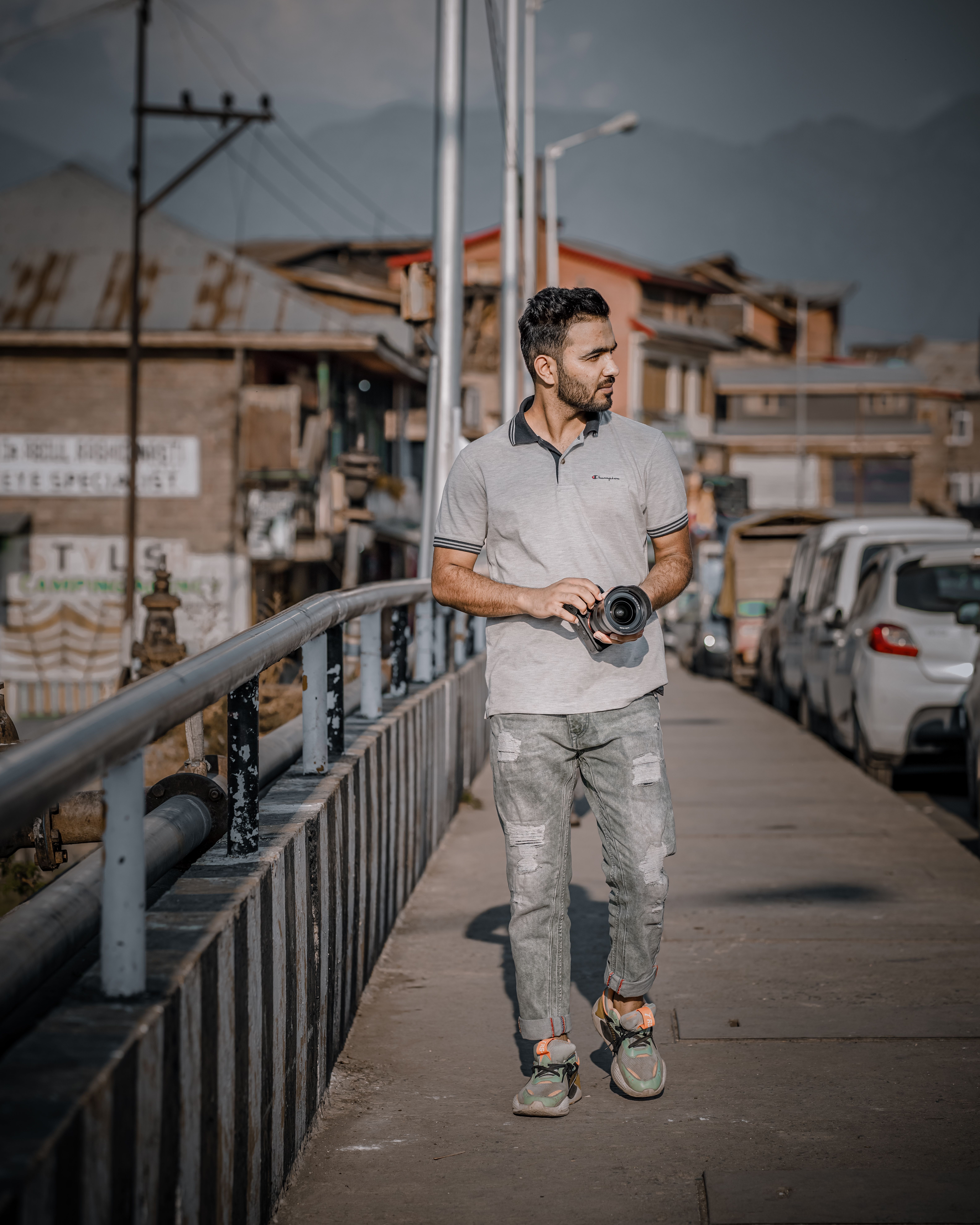 A man holding a camera walks on a sidewalk in a lively urban street scene with mountains in the background.