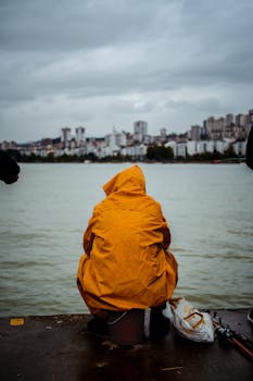 A man in a yellow raincoat fishes by the seaside under overcast skies, city skyline in the background.