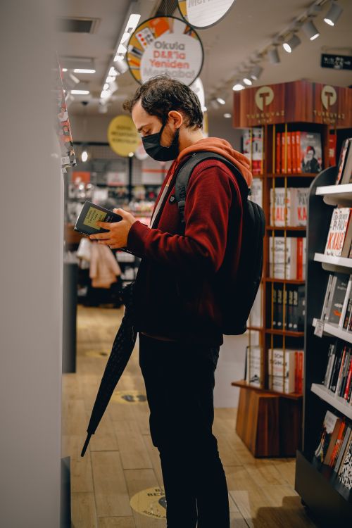 Free A man wearing a face mask reads a book in a lively bookstore setting. Stock Photo