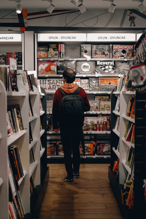 Free Man with a backpack browsing board games in a store aisle filled with books and games. Stock Photo