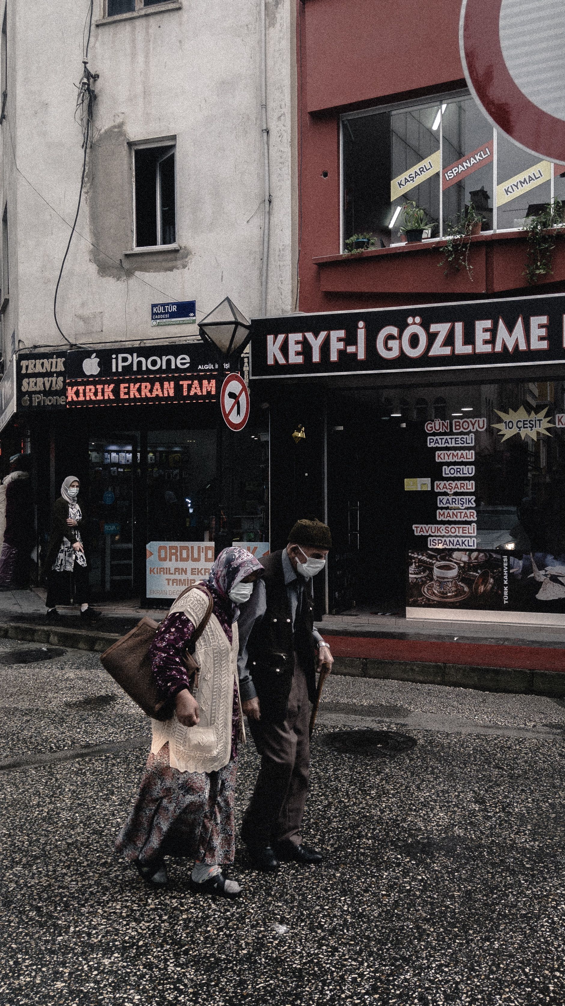 An elderly couple walks past storefronts on a wet city street, both wearing masks.