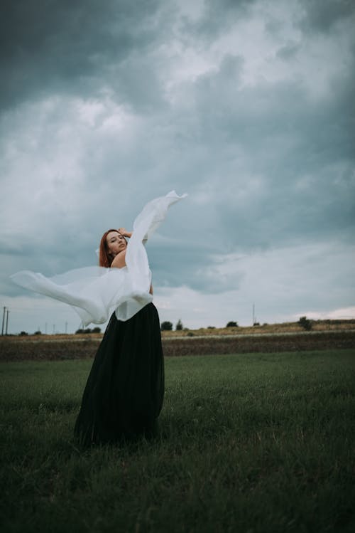 Free Woman in White Long Sleeve Shirt and Black Skirt Standing on Green Grass Field Under White Stock Photo