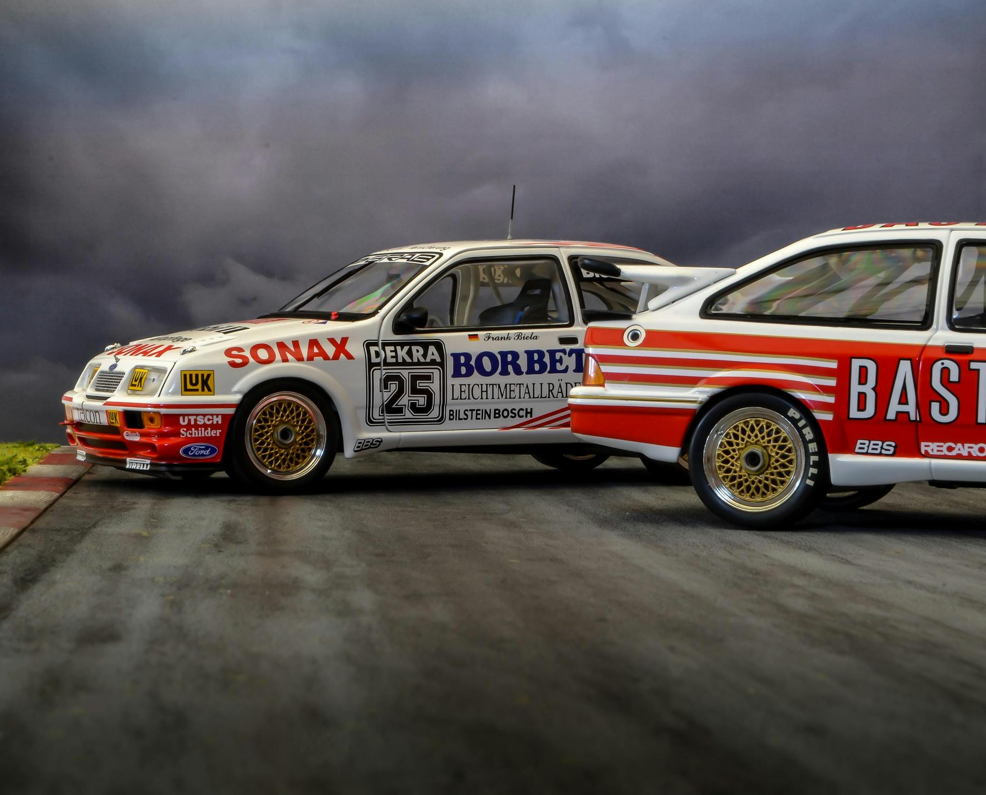 Close-up shot of classic motorsport cars on a racing track against a dramatic cloudy backdrop.