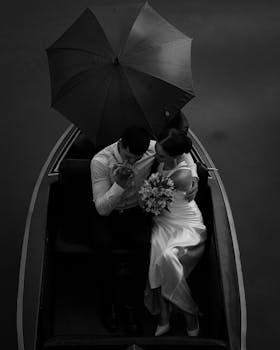 Black and white photo of a wedding couple in a boat holding an umbrella, embracing a romantic moment.