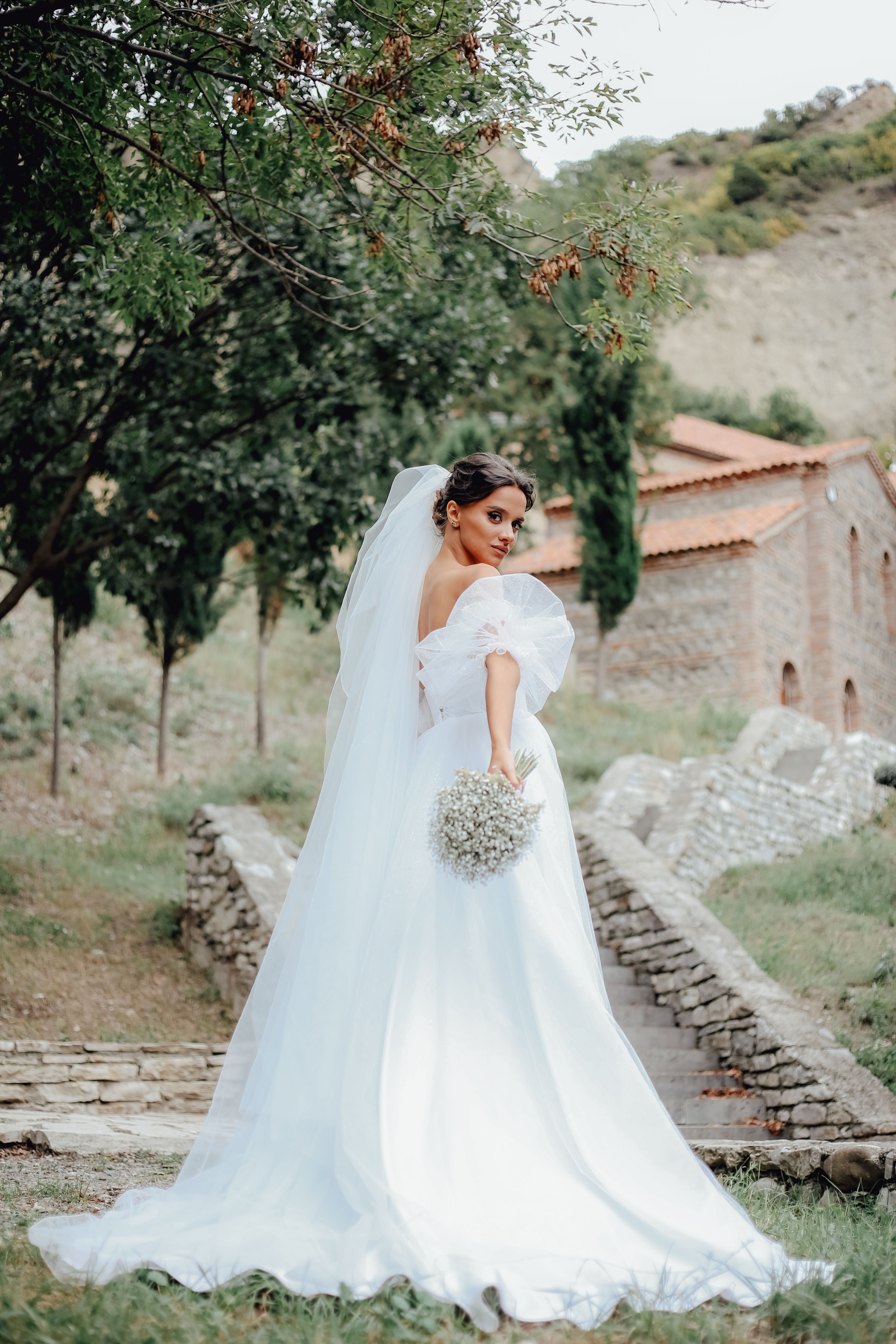 Bride in a white wedding dress holding a bouquet, posing outdoors in a scenic location.