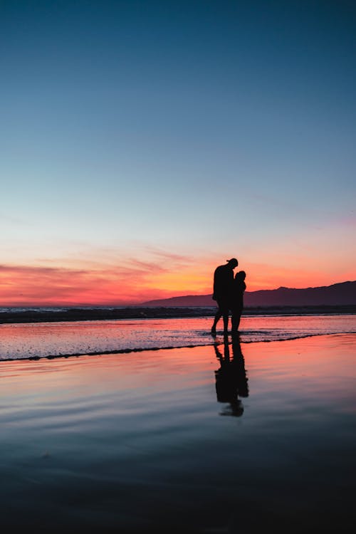 Free A Silhouette of a Couple Standing on Seashore during Sunset Stock Photo