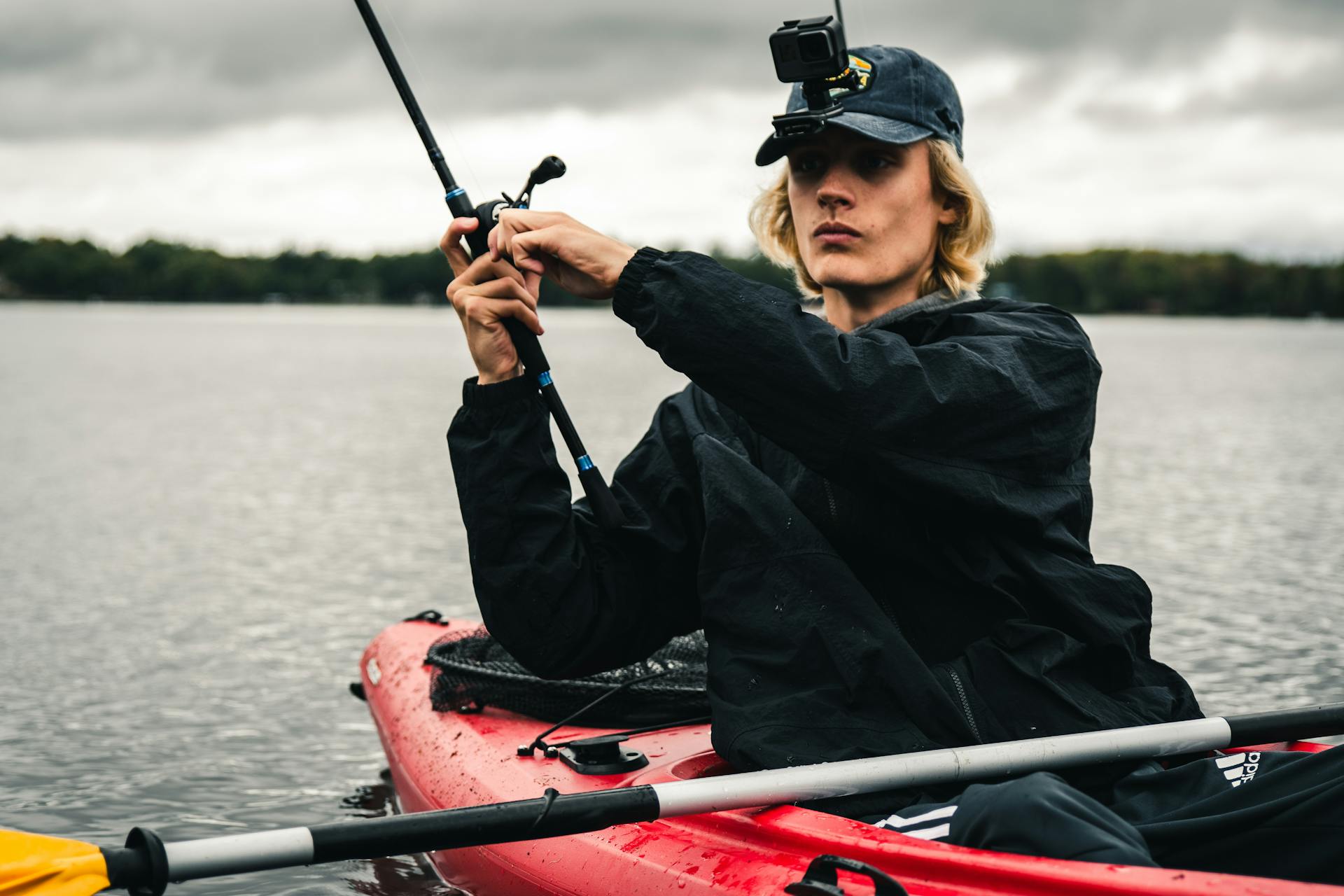 A man fishing from a kayak wearing a cap and GoPro on a calm lake.