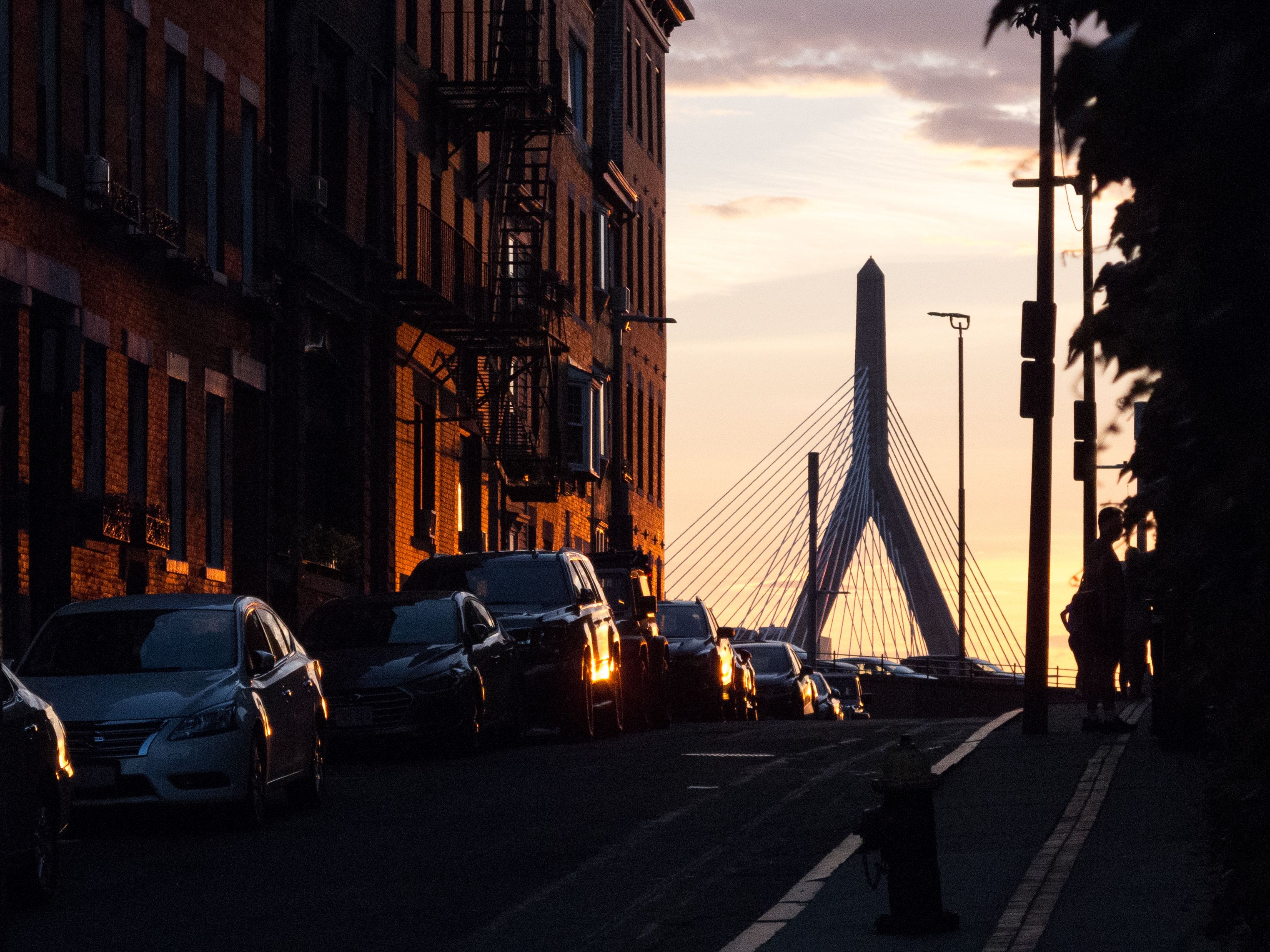 Cars parked along a Boston street silhouetted against the sunset with Zakim Bridge in the background.