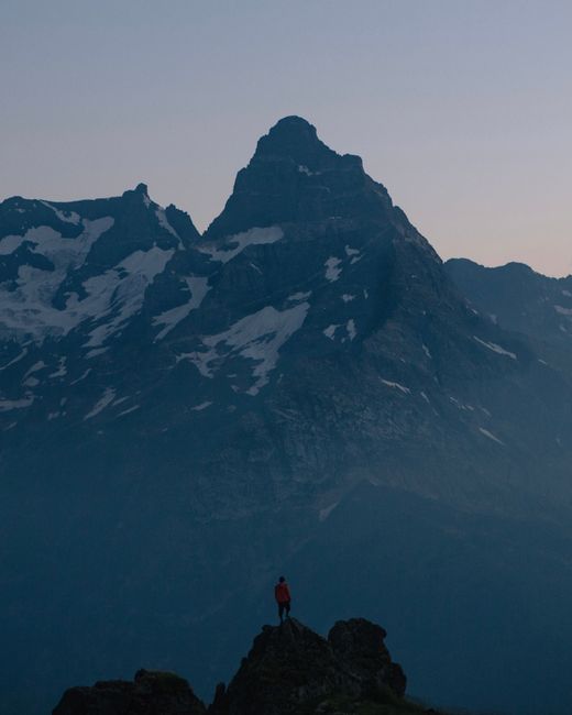 A lone hiker stands atop a rocky peak against a stunning mountain backdrop at dusk.