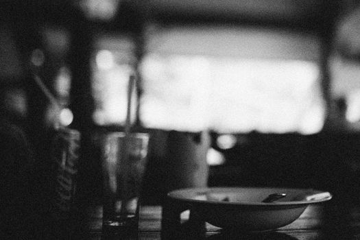 Black and white image showing a table setting with focus on a glass and a plate in a dimly lit environment.