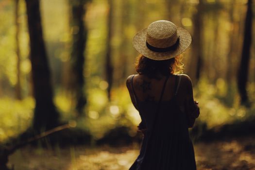 A woman with tattoos and straw hat stands in a sunlit forest, embracing nature.