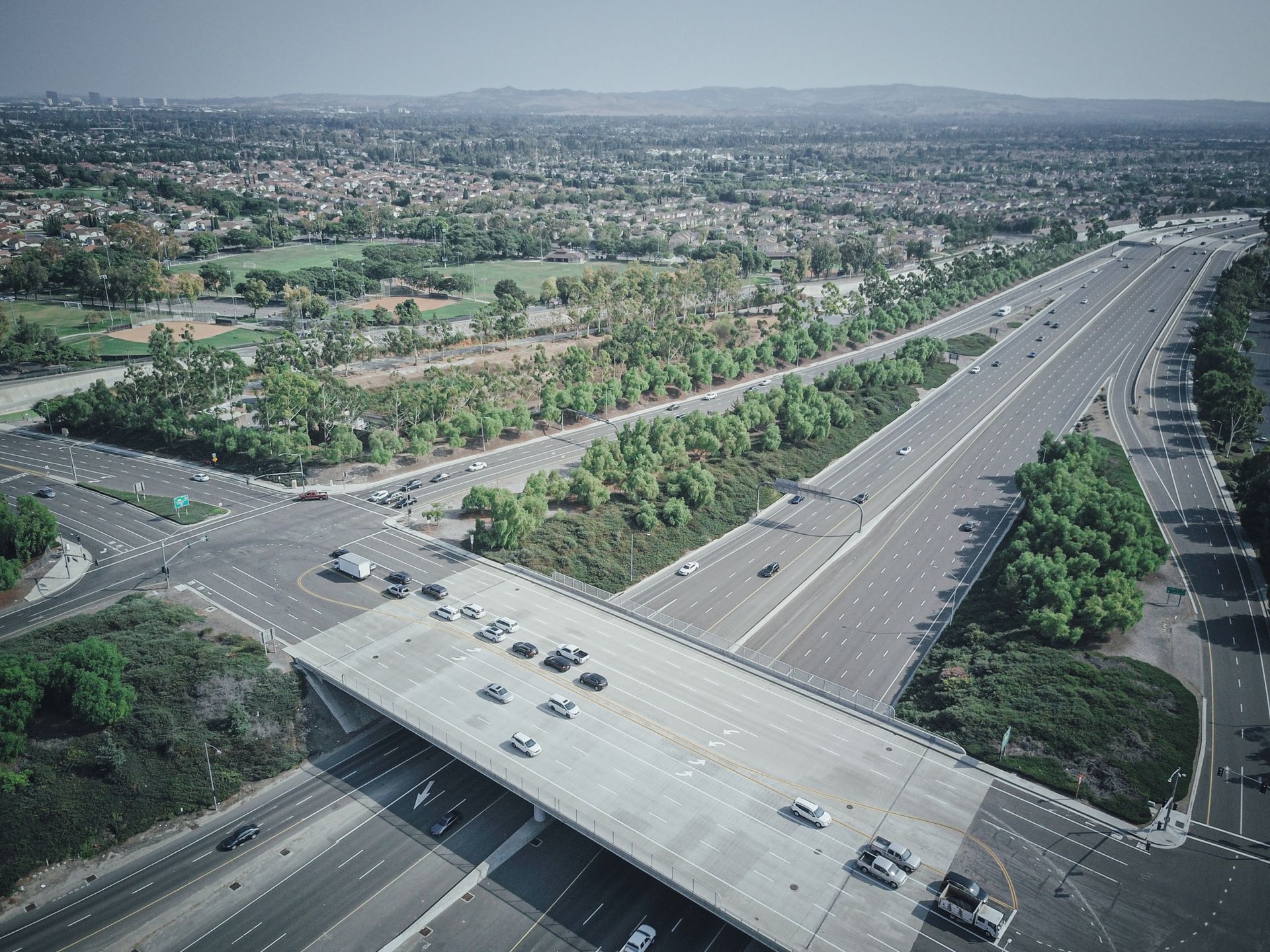 Drone view of cars on a wide highway intersection surrounded by greenery in an urban setting.