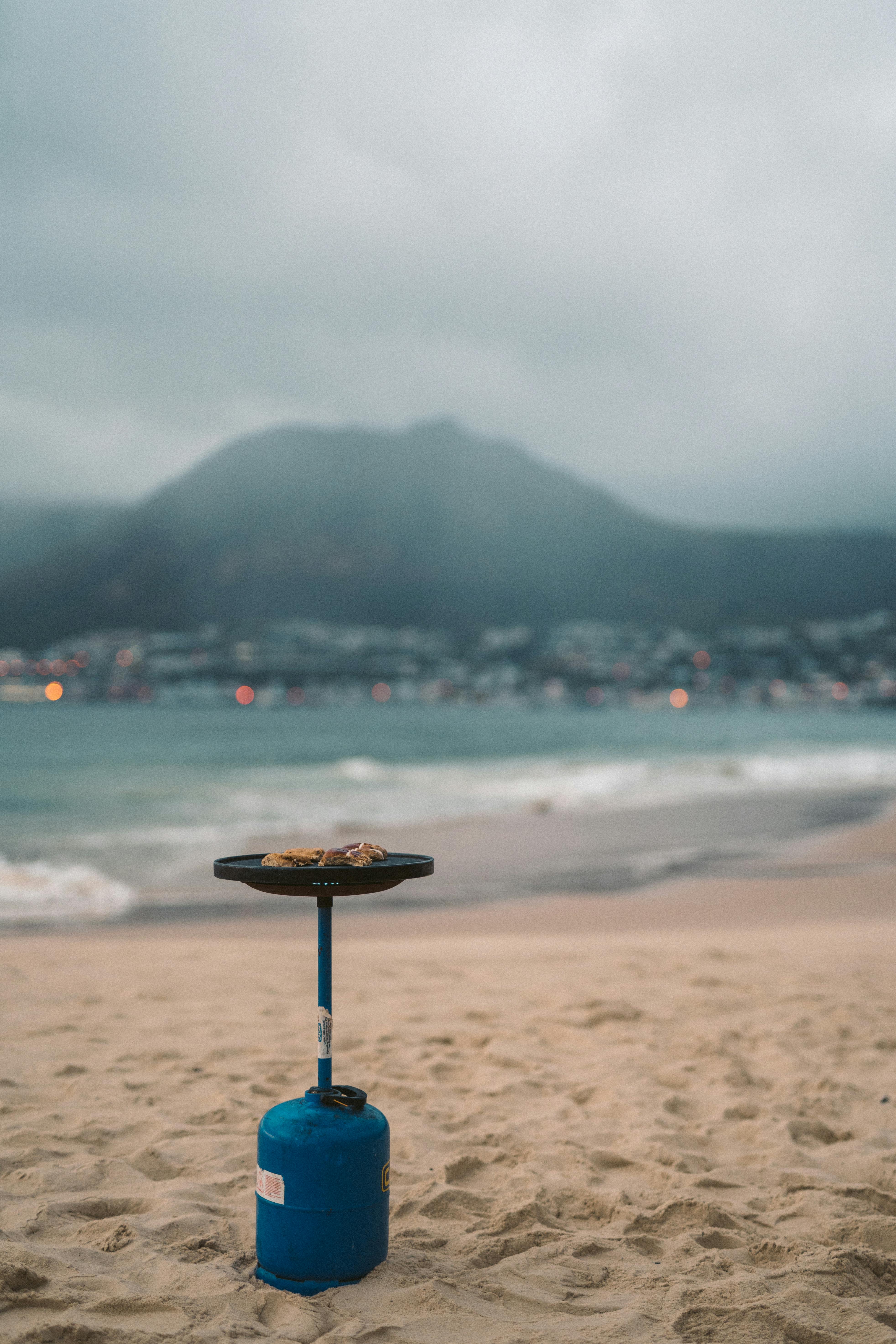 A portable gas bottle grill setup on a sandy beach with a misty mountain backdrop.