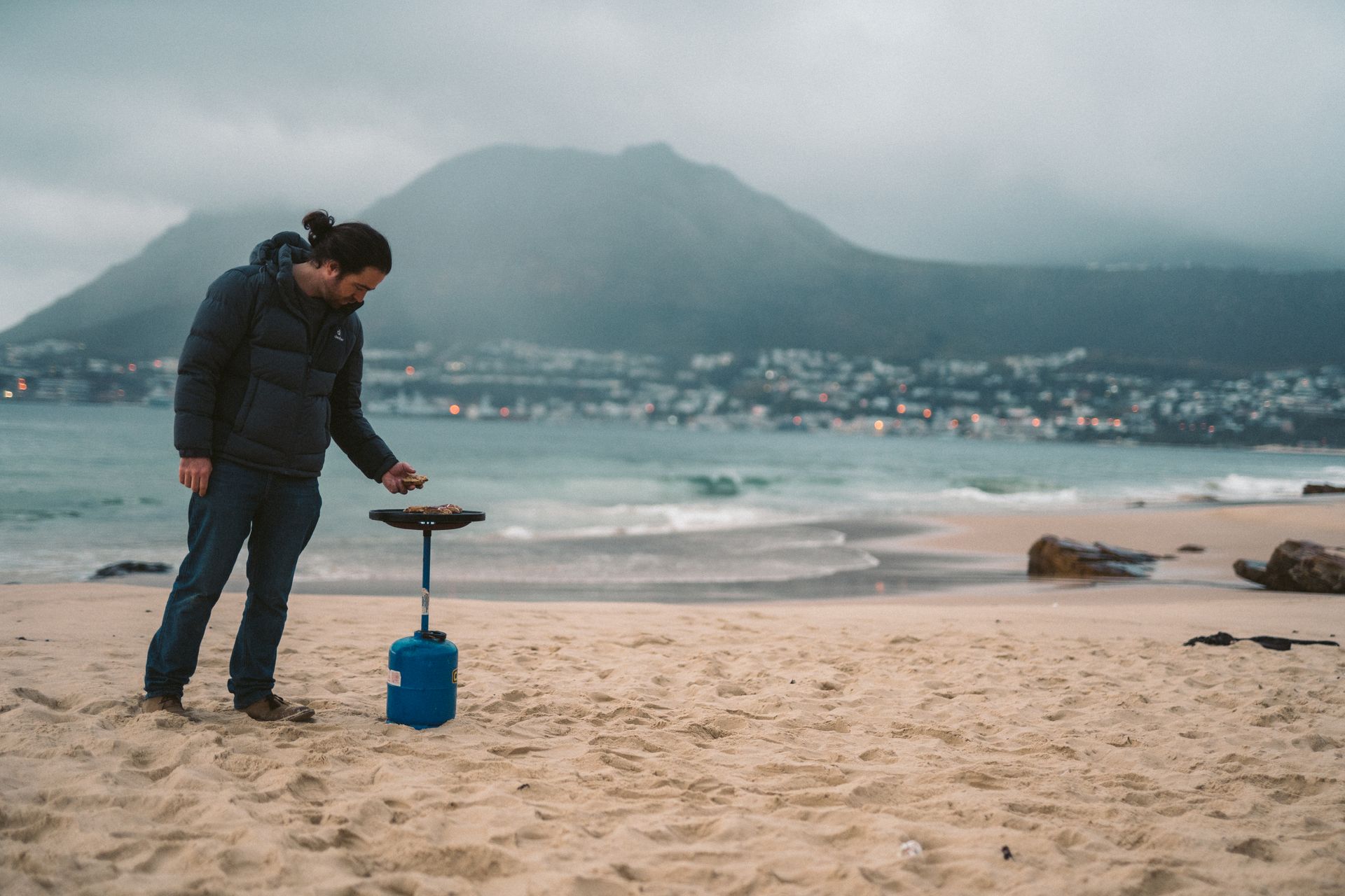 A man cooks on a portable gas stove on a sandy beach with ocean waves and mountains in the background.