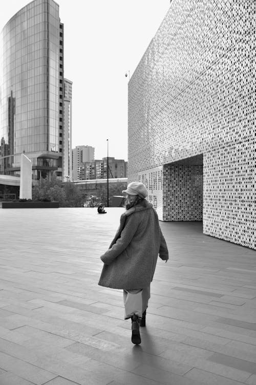 Grayscale Photo of a Woman in a Coat Walking at a City