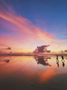 Dramatic sunset reflections on a beach with silhouettes of two people walking.