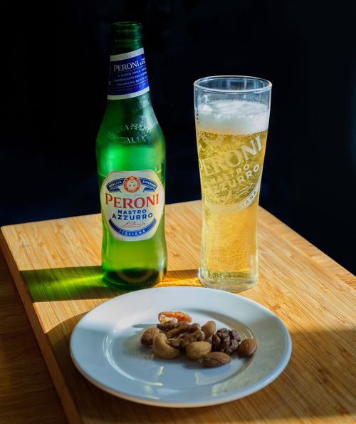 Free Peroni Nastro Azzurro beer paired with a plate of assorted nuts on a wooden table. Stock Photo