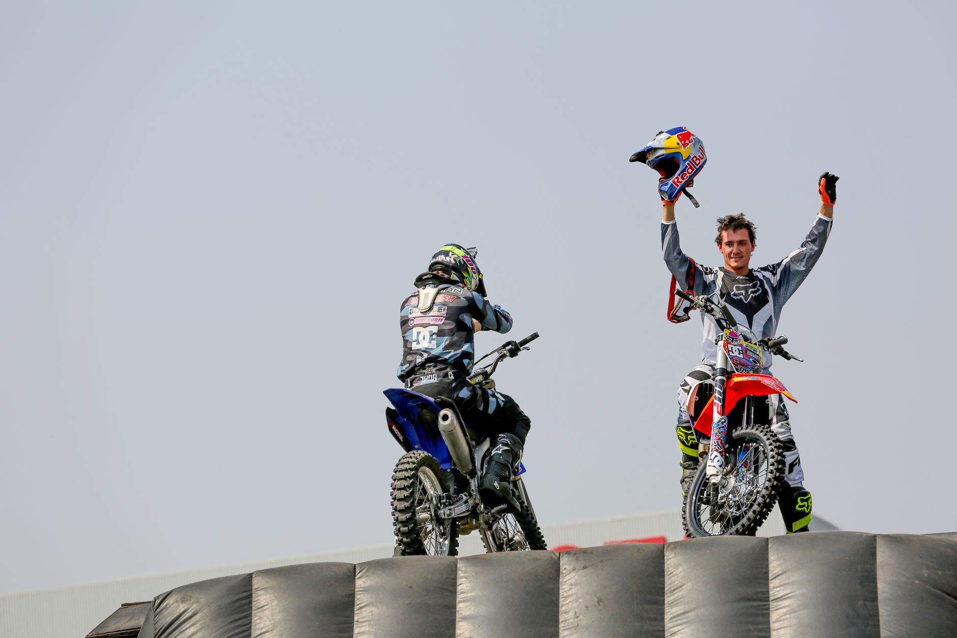 Two motocross riders on bikes, one raising a helmet victoriously outdoors.