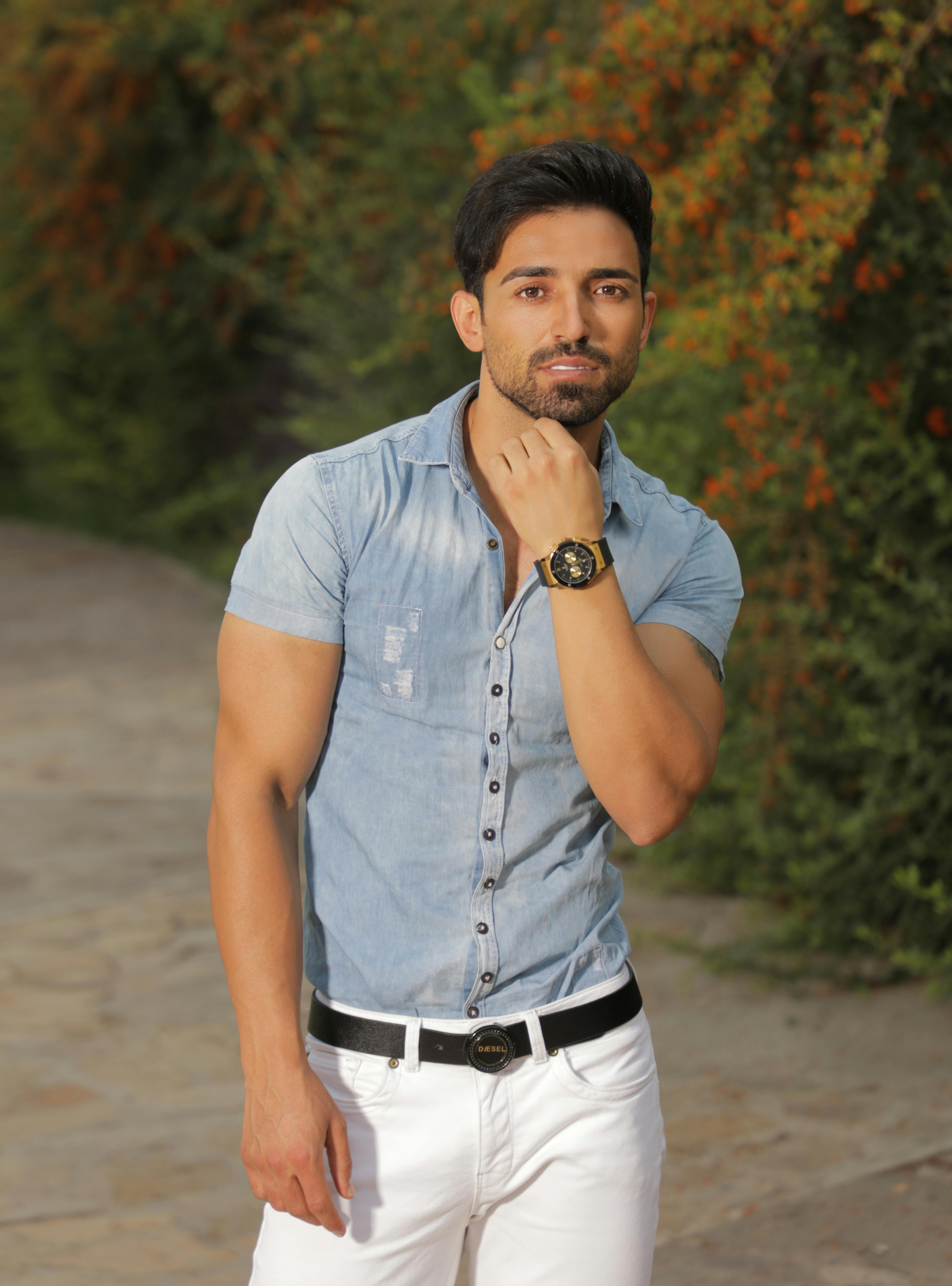 A stylish man poses outdoors in a denim shirt with lush greenery background.