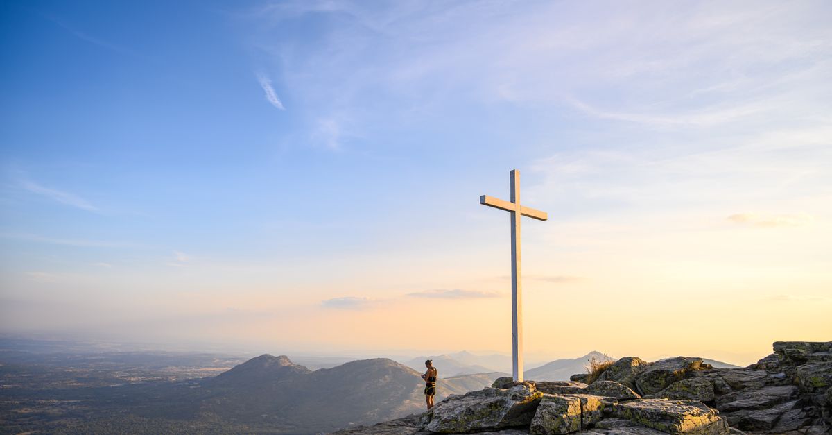A hiker stands by a mountain cross at sunrise in Spain, surrounded by scenic vistas and serene skies.