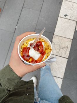 A person holds a cup of corn topped with ketchup and mayonnaise, photographed from above.
