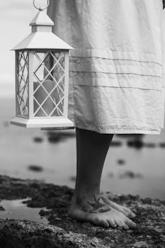 A grayscale photo of a woman holding a lantern by the sea, capturing tranquility.