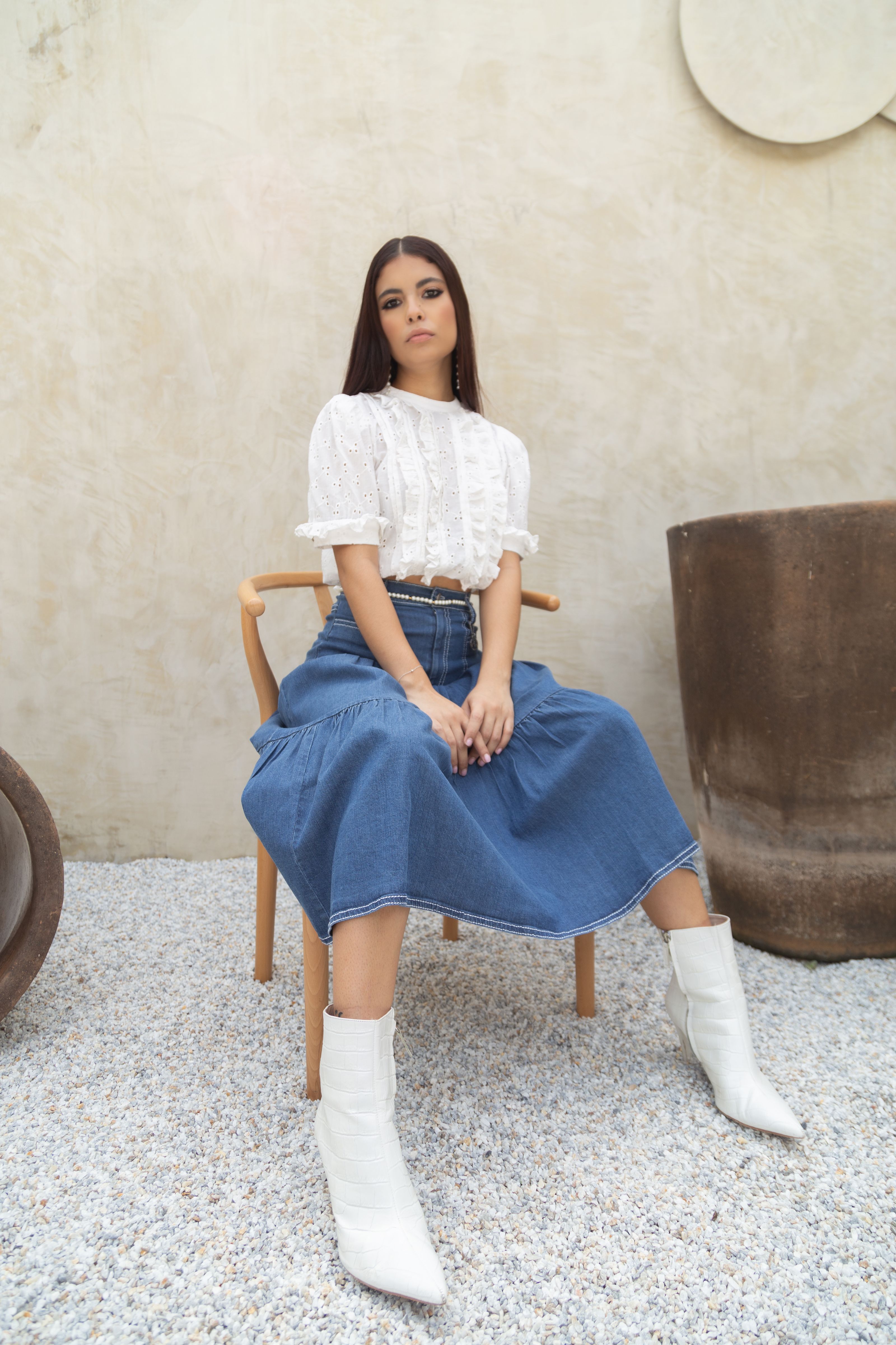 Mulher estilosa sentada em uma cadeira com saia jeans, blusa branca e botas.