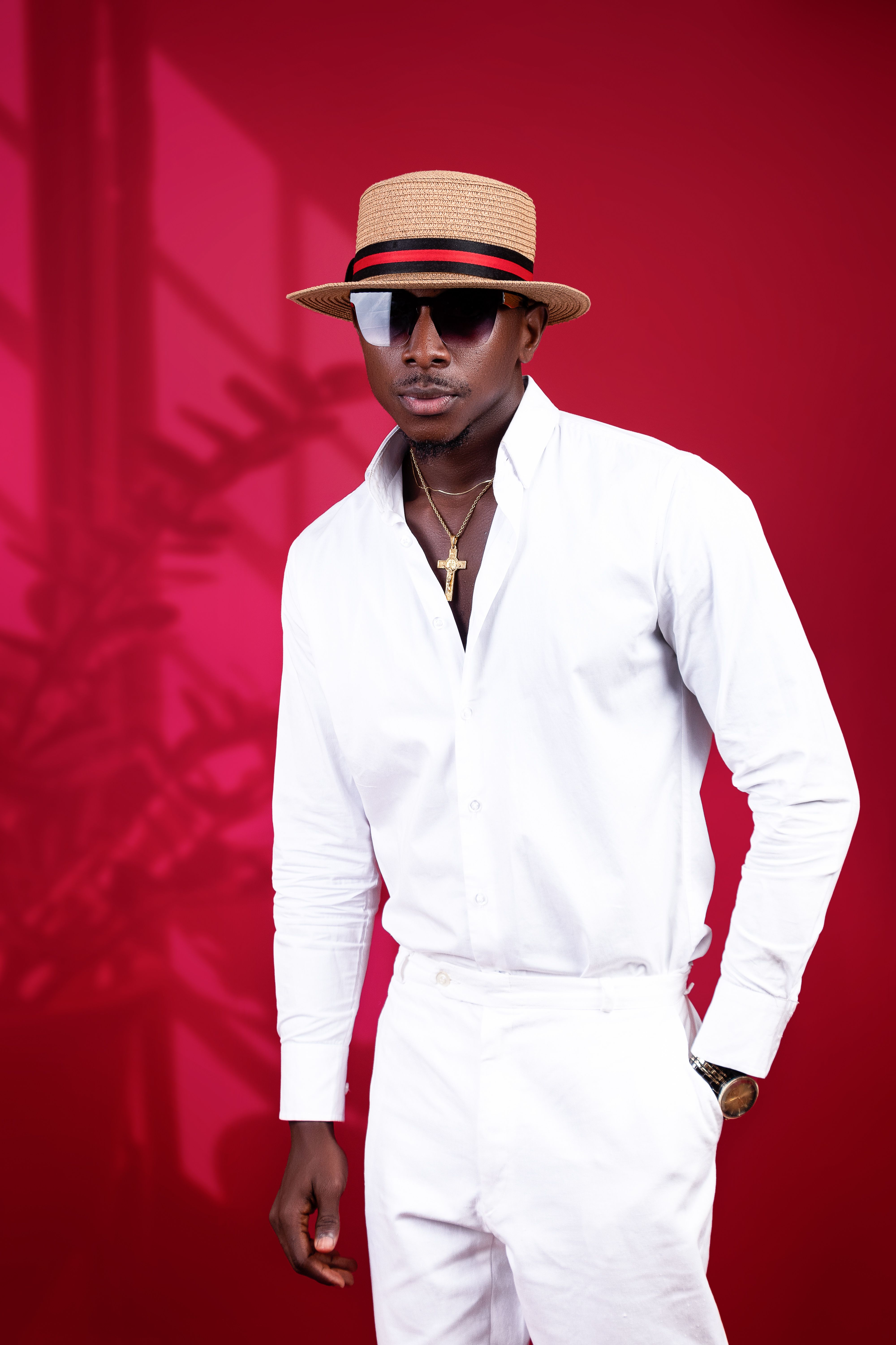Fashionable man posing in white attire and straw hat against red background.