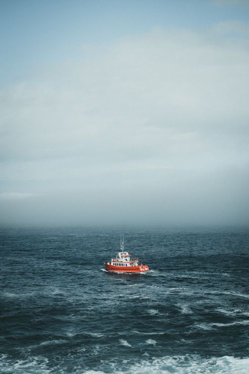Free A solitary red boat navigating through a vast blue ocean with an expansive cloudy sky above. Stock Photo