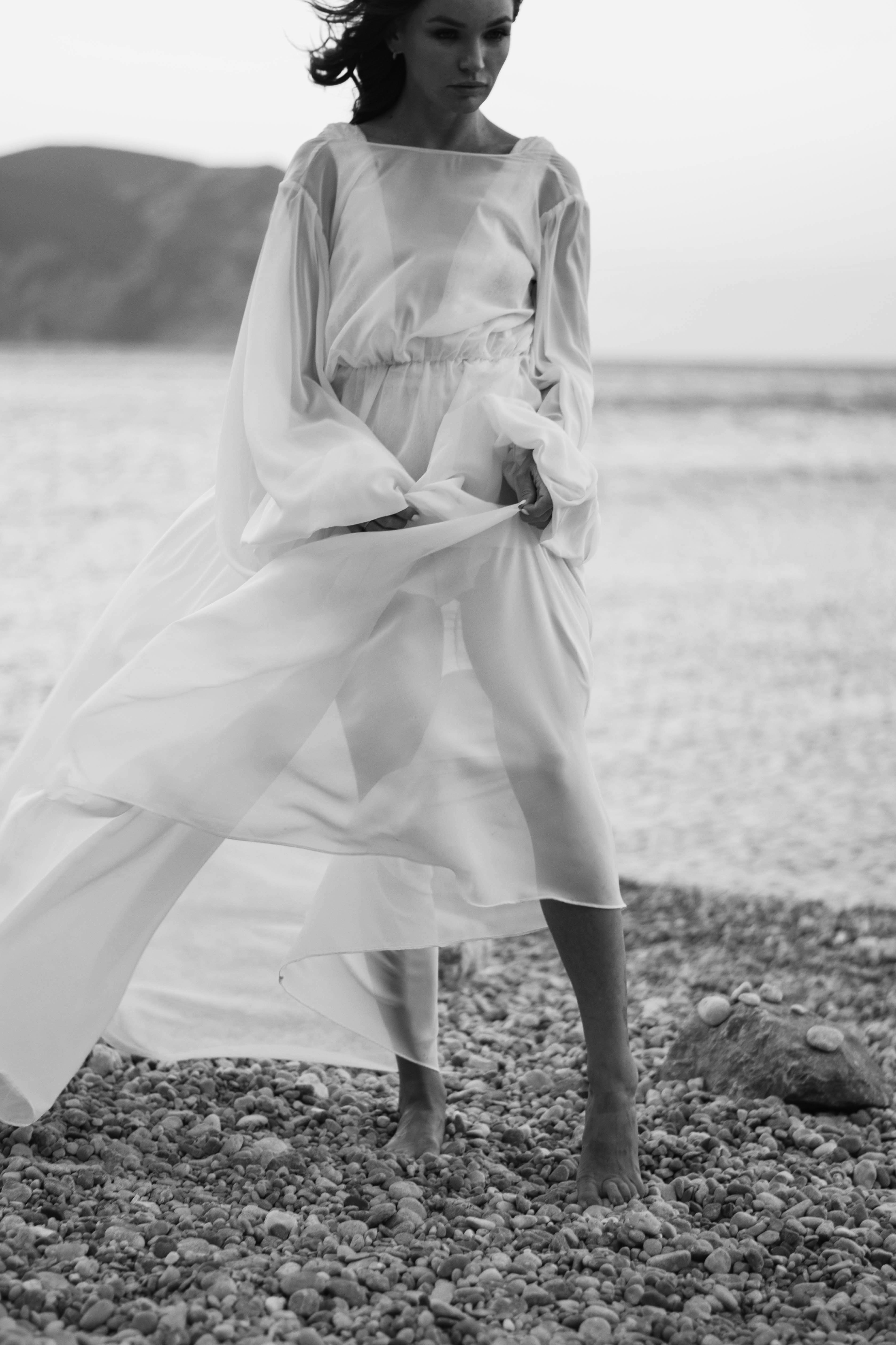 A woman in a flowing dress stands barefoot on a pebble beach, captured in striking black and white.