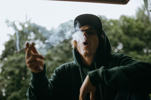 Free A young man in a hoodie smoking outdoors, exhaling smoke with trees in the background. Stock Photo