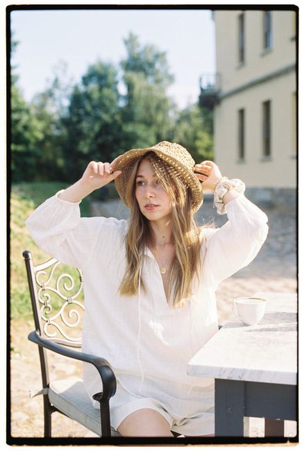 A woman in a sun hat and white dress seated outdoors at a table, enjoying a sunny day.