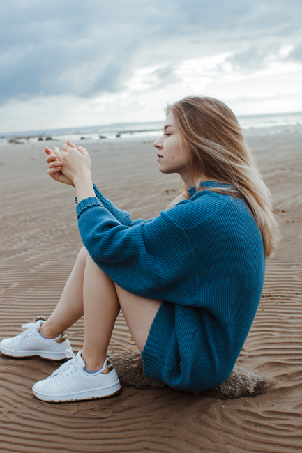 A woman in a blue sweater sits pensively on a sandy beach with overcast skies.