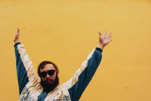 A bearded man wearing sunglasses poses with arms raised against a vibrant yellow wall.