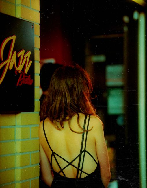 A woman in a black dress stands near a jazz club entrance with a lit sign.